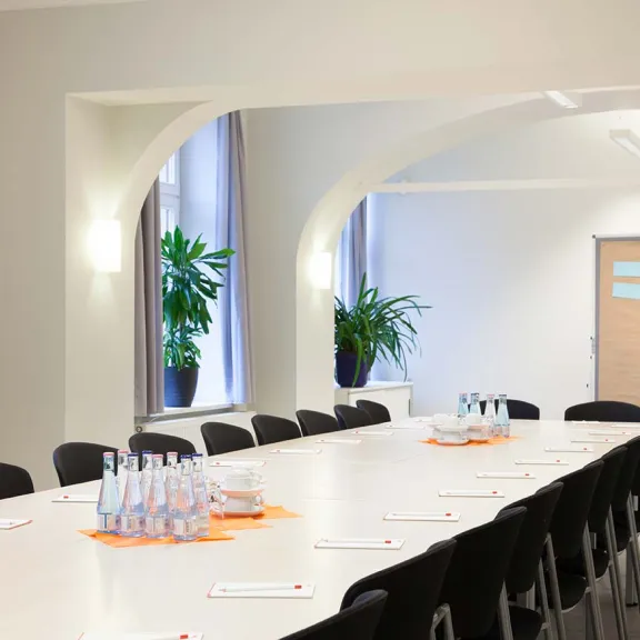 Heller Konferenzraum mit langem Tisch, Stühlen, Wasserflaschen und Flipchart.