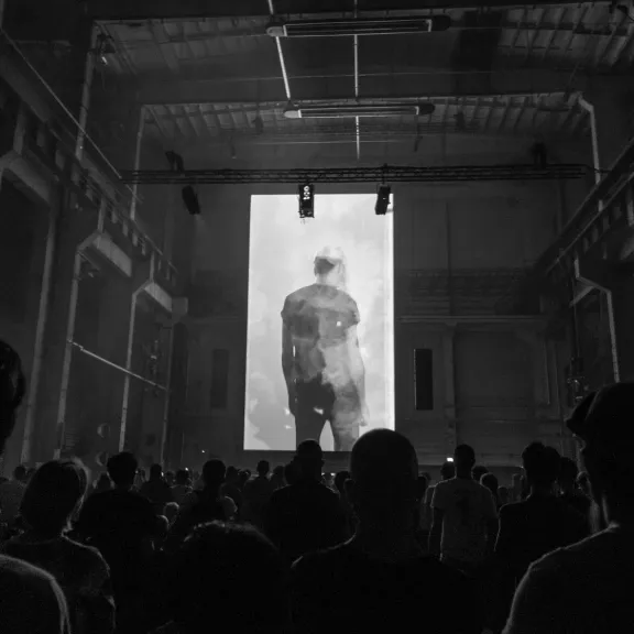 Menschenmenge blickt auf große Leinwand in dunkler Industriehalle.