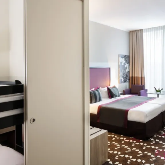 Modernes Hotelzimmer mit Doppelbett, Etagenbett und zwei lila Sesseln am Fenster.
