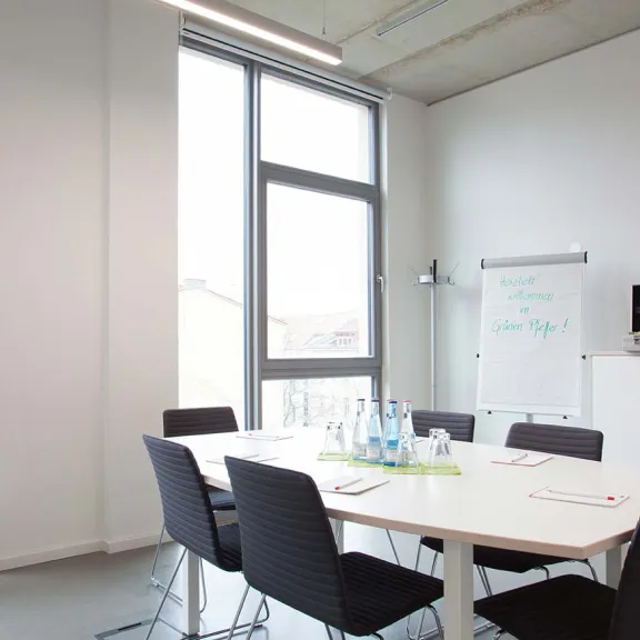Heller Konferenzraum mit Tisch, sechs Stühlen und Flipchart neben großen Fenstern.