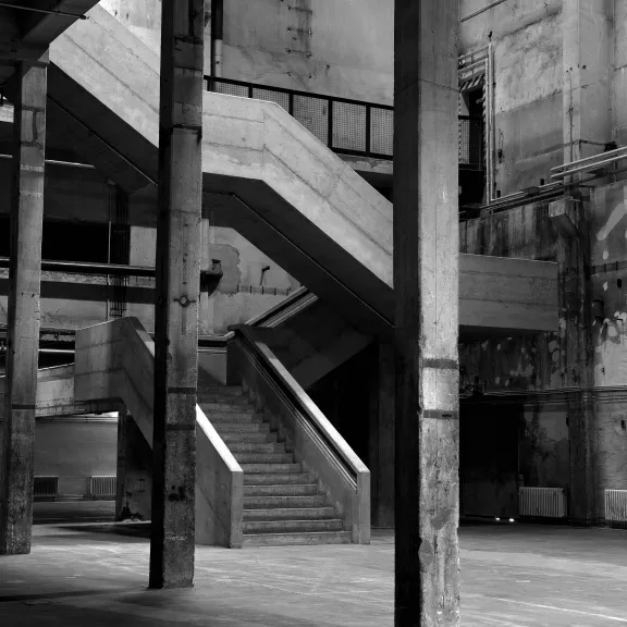 Alte, leere Fabrikhalle mit Treppe, rauen Wänden und Säulen in Schwarz-Weiß.