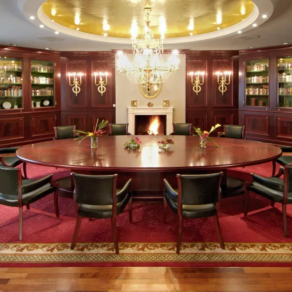 Edler Konferenzraum mit ovalem Holztisch, Kamin, Kronleuchter und roten Teppichen.
