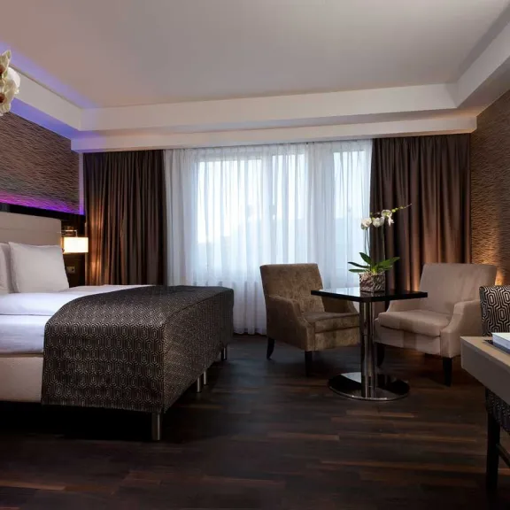 Modernes Hotelzimmer mit Doppelbett, zwei Sesseln, Tisch und großen Fenstern mit Vorhängen.