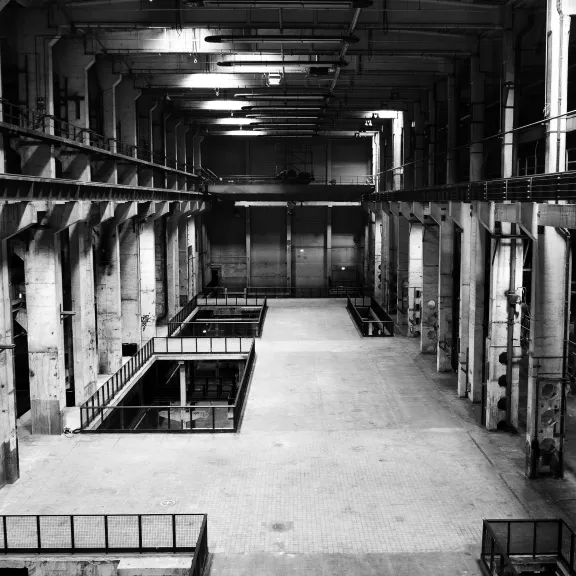 Große, leere Industriehalle mit Betonwänden und hohen Decken in Schwarzweiß.