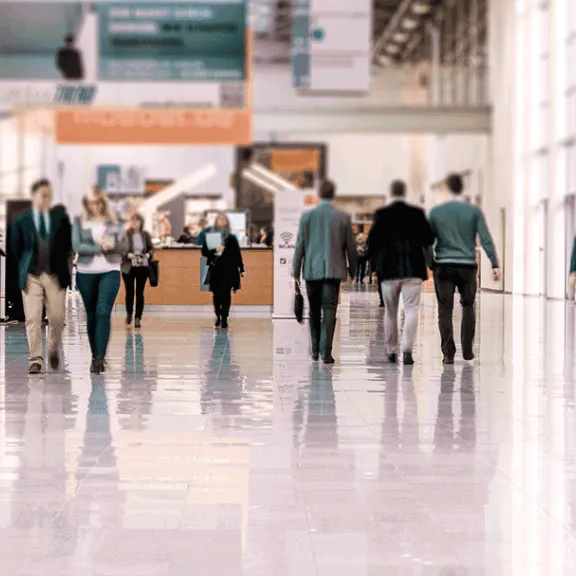 Menschen gehen durch eine helle, moderne Halle mit Glasfronten und glänzendem Boden.