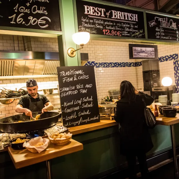 Imbissstand mit Koch, der Essen zubereitet, Gäste stehen an der Theke.