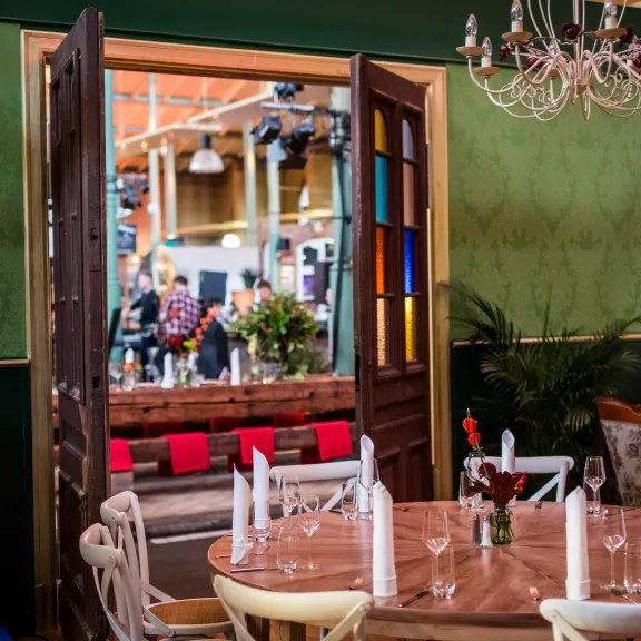 Rund gedeckter Tisch in grünem Raum, offene Tür zeigt belebte Halle im Hintergrund.