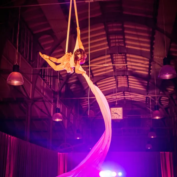 Akrobatin schwebt an langen Tüchern unter hallenartigem Dach, lila Licht im Hintergrund.