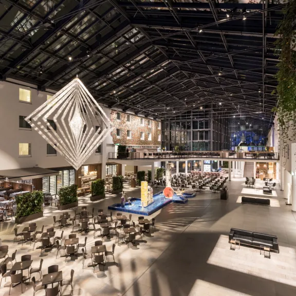 Große, moderne Hotel-Lobby mit Glasdach, vielen Tischen und geometrischer Skulptur.