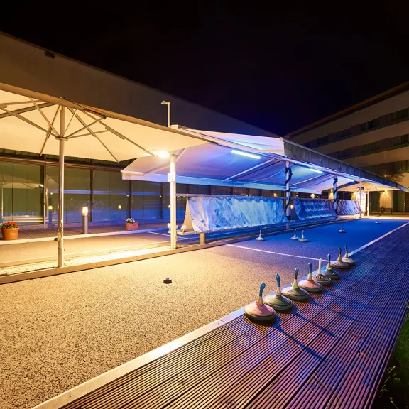 Beleuchtete Terrasse mit Sonnenschutz, mehreren Entenskulpturen und modernen Gebäuden bei Nacht.