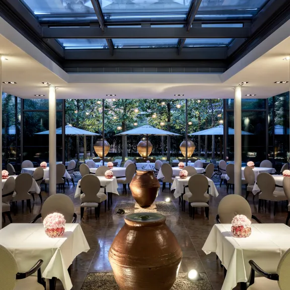 Modernes Restaurant mit weißen Tischen, großen Fenstern und Ausblick auf einen beleuchteten Garten.