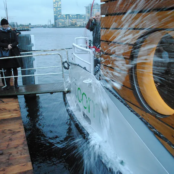 Eine Frau zerschlägt eine Sektflasche an einem Schiffsrumpf bei einer Taufe.