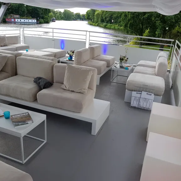 Moderne Lounge mit hellen Sofas auf einem Boot, Ausblick auf einen Fluss und grüne Bäume.