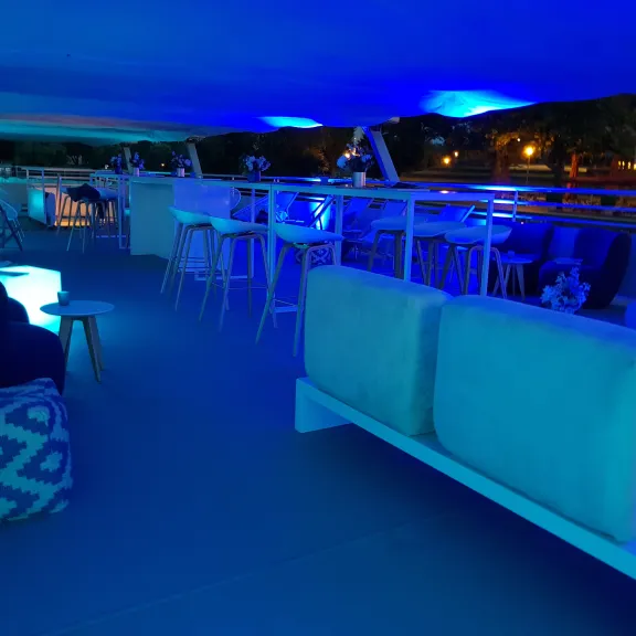 Lounge mit blauen Lichtern, modernen Sofas und hohen Tischen auf einer Terrasse bei Nacht.