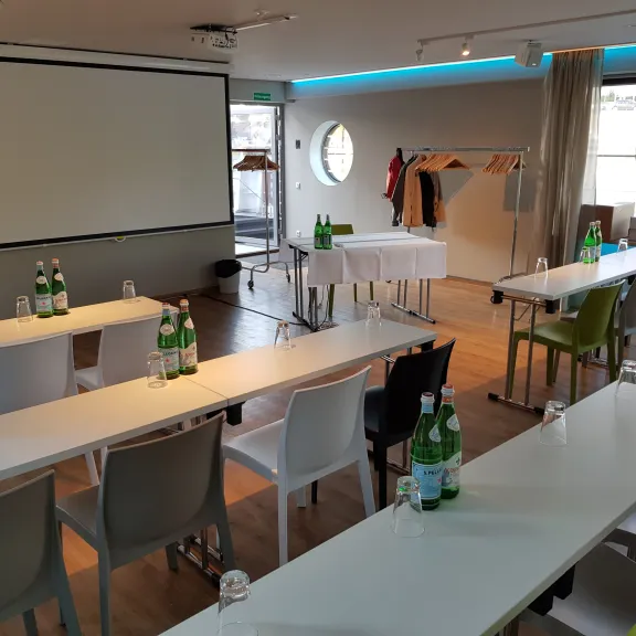 Konferenzraum mit Tischen, Stühlen, Wasserflaschen und großem Projektor.