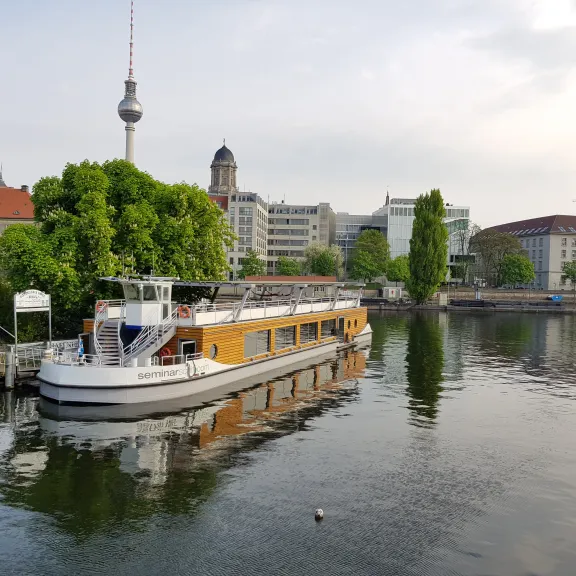 Ausflugsschiff auf einem Fluss in Berlin mit Fernsehturm und grünen Bäumen im Hintergrund.