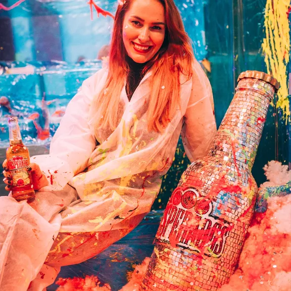 Frau in weißem Kittel lächelt, hält Getränk, neben großer glitzernder Flasche.