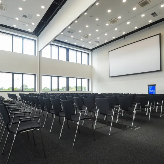 Großer, leerer Konferenzraum mit vielen schwarzen Stühlen und großer Leinwand.