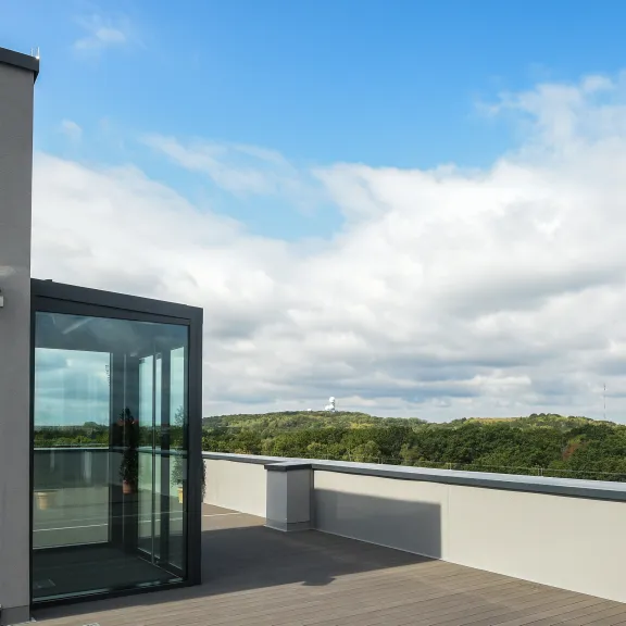 Moderne Dachterrasse mit Glasaufzug, blauer Himmel und grüne Bäume im Hintergrund.
