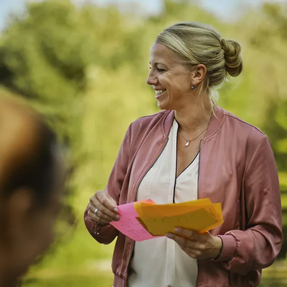Frau mit rosa Jacke hält bunte Zettel und lächelt im Grünen.