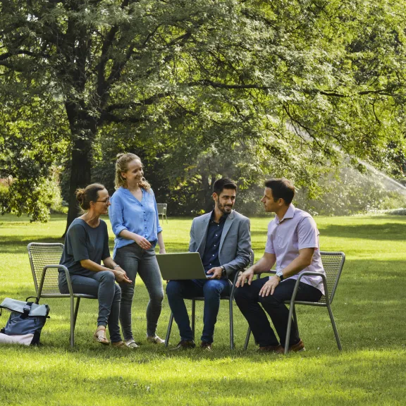 Vier Personen sitzen im Park auf Stühlen und unterhalten sich, Laptop auf dem Tisch.