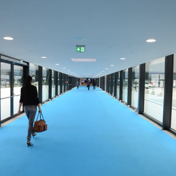 Frau mit Tasche geht auf blauem Teppich in heller Glasgang-Brücke.