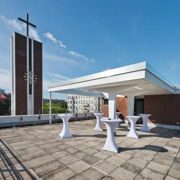 Dachterrasse mit Stehtischen, im Hintergrund ein Kirchturm mit Kreuz bei blauem Himmel.