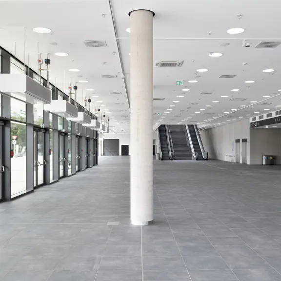 Heller, leerer Eingangsbereich mit Glasfront, Säule und Rolltreppen im Hintergrund.