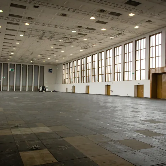 Großer leerer Saal mit vielen Fenstern und Deckenleuchten, Tageslicht strömt herein.