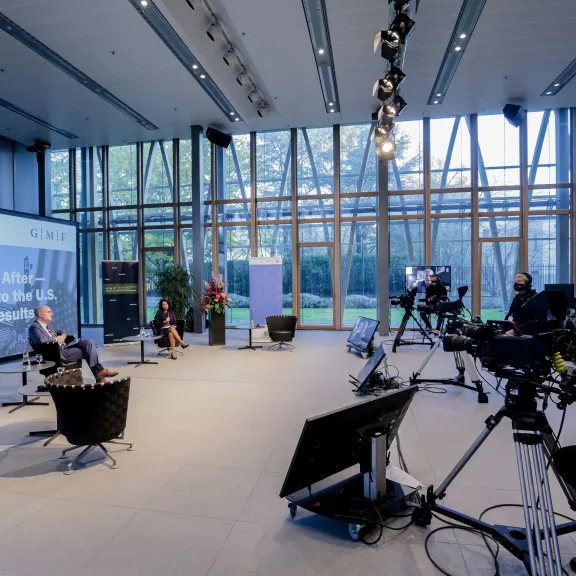 Konferenzraum mit Kameraausrüstung, drei Personen diskutieren vor großer Fensterfront.