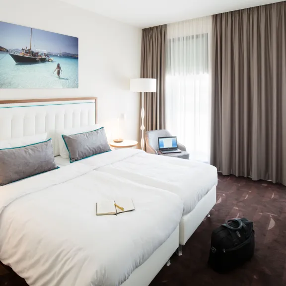 Modernes Hotelzimmer mit Doppelbett, großem Fenster, Schreibtisch und Reisetasche am Boden.