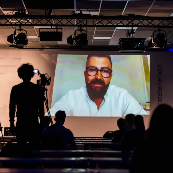 Großer Bildschirm mit Videokonferenz, Zuschauer sitzen im dunklen Konferenzraum.