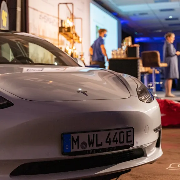 Weißer Tesla mit deutschem Kennzeichen im Vordergrund, Bühne mit Menschen im Hintergrund.