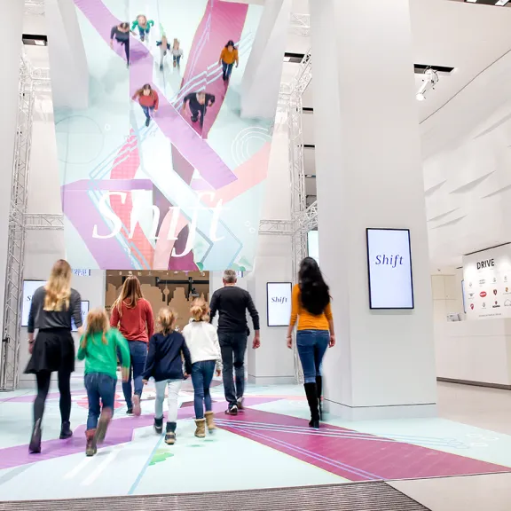 Mehrere Menschen betreten eine moderne, helle Einkaufspassage mit buntem Bodenmuster.