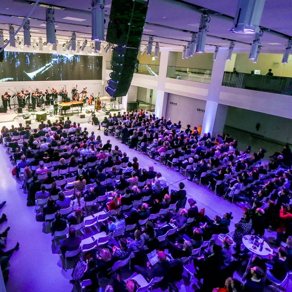 Großer Konzertsaal mit Publikum, Bühne und Musikern, beleuchtet in lila Licht.