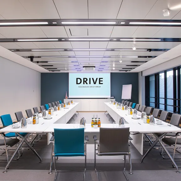 Moderner Konferenzraum mit U-förmigem Tisch, Stühlen und Bildschirm mit Schriftzug "DRIVE".