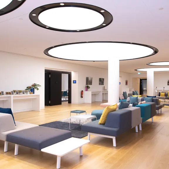 Moderner Wartebereich mit bunten Sofas, Holzboden und großen runden Deckenleuchten.