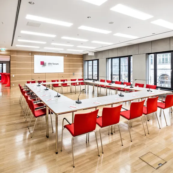 Moderner Konferenzraum mit roten Stühlen, U-förmig angeordnet, große Fenster, helle Beleuchtung.