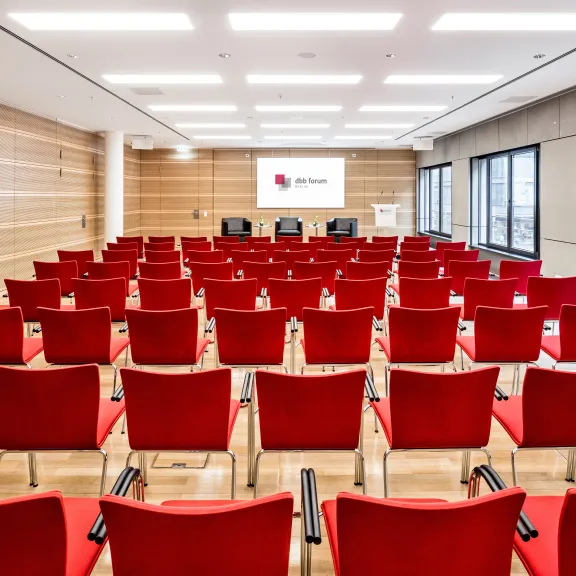 Konferenzraum mit vielen roten Stühlen und großer Fensterfront, vorne ein Podium.