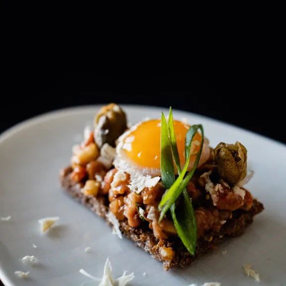 Dunkles Brot mit Gemüse, Spiegelei und grüner Garnitur auf weißem Teller.