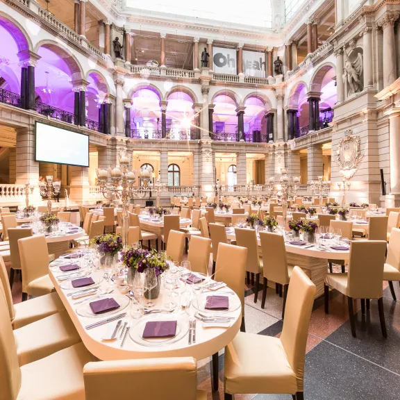 Festlich gedeckter Saal mit runden Tischen, lila Beleuchtung und elegantem Ambiente.