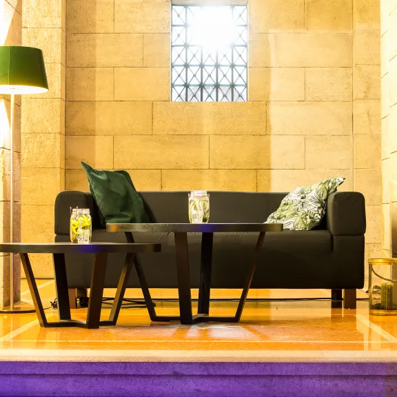 Modernes Wohnzimmer mit dunklem Sofa, Kissen, zwei Tischen und Stehlampe vor Steinwand.