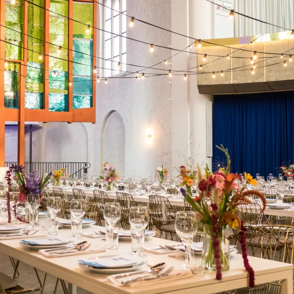 Festlich gedeckte Tafeln mit Blumen, Lichterketten und bunten Fenstern in hellen Saal.