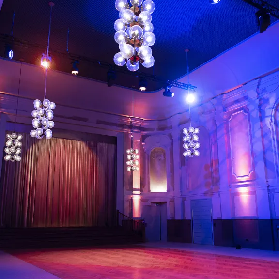 Der große Festsaal des Peter Edel mit Effektbeleuchtung, man sieht den Blick auf die Bühne, einen Bühnenvorhang sowie die neobarocken Zierelemente an den Wänden und die Deckenlampen, die im Stil des Palastes der Republik gehalten sind