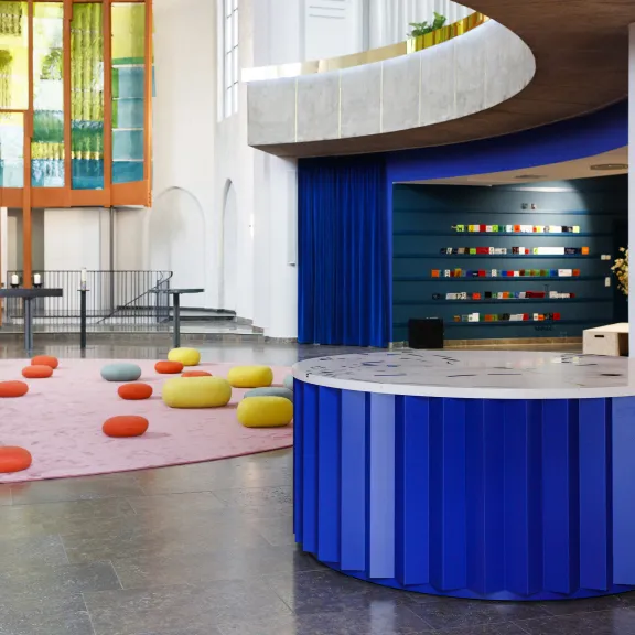 Moderner, heller Raum mit bunten Sitzkissen, rundem blauen Tresen und großem Fenster.