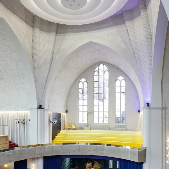 Heller Kirchenraum mit moderner Einrichtung, gelbe Sitzbank vor hohen Fenstern.