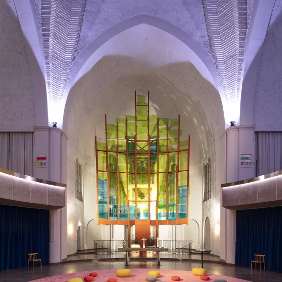 Großer Saal mit hoher Decke, bunten Kissen und moderner Glasinstallation.