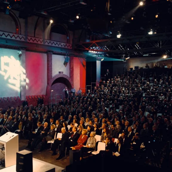 Großer Saal mit Bühne, Redner am Podium, viele Menschen im Publikum.