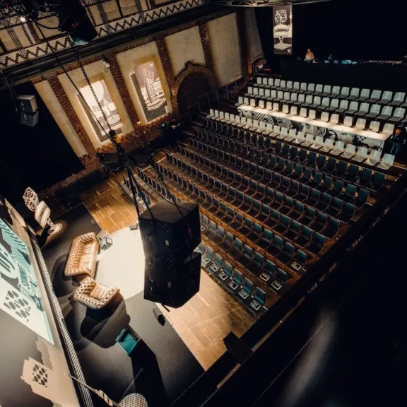 Leerer Veranstaltungssaal mit Bühne, Sesseln und Reihen von Stühlen.