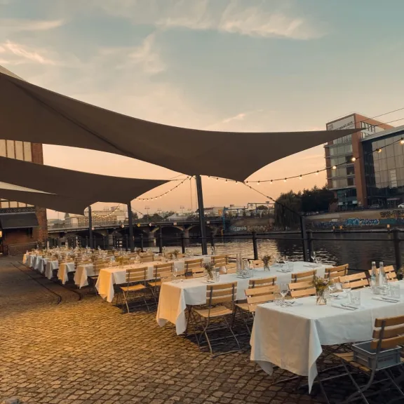 Lange, festlich gedeckte Tische draußen am Wasser bei Sonnenuntergang.